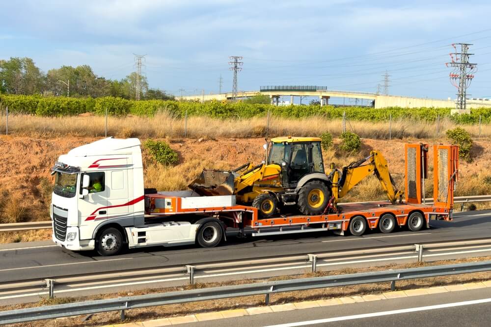 Biały ciężarowy ciągnik transportujący żółto-czarną maszynę budowlaną na przyczepie, poruszający się po drodze w otoczeniu zielonych terenów.